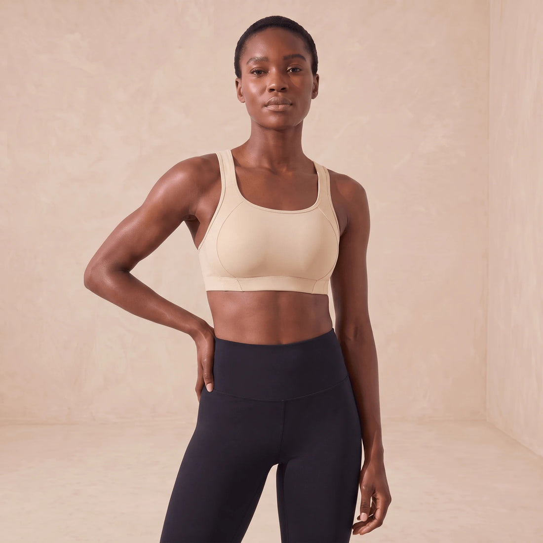 Femme en brassière de sport beige à maintien élevé, vue de face, mains sur les hanches, bretelles larges