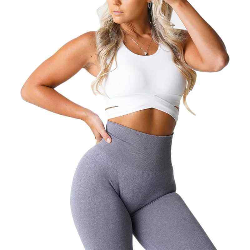 Brassière sport femme sans couture avec croisement de bretelles, portée avec leggings gris, crop top blanc.