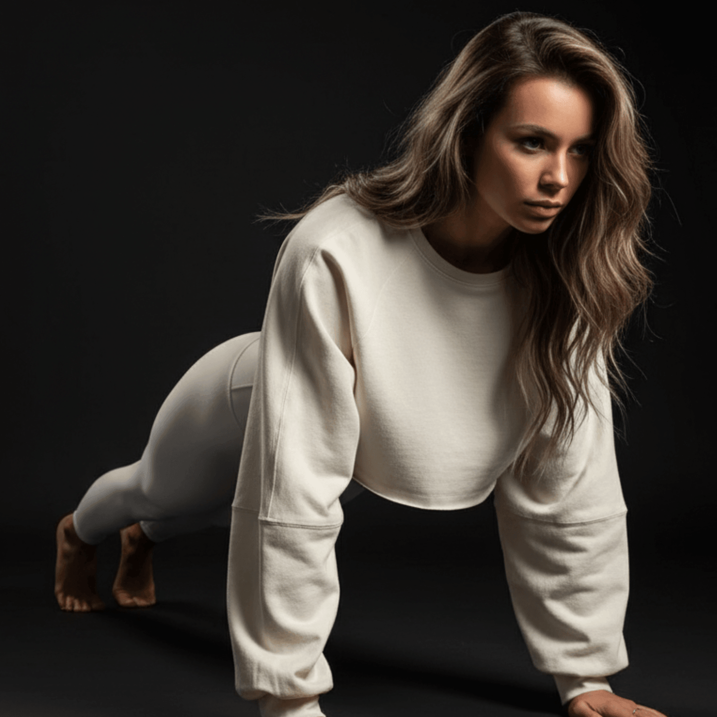 Femme en tenue de sport blanche, sweat-shirt oversized et pantalon assorti, accroupie sur le sol. Cropped top femme sport visible, ambiance sombre.