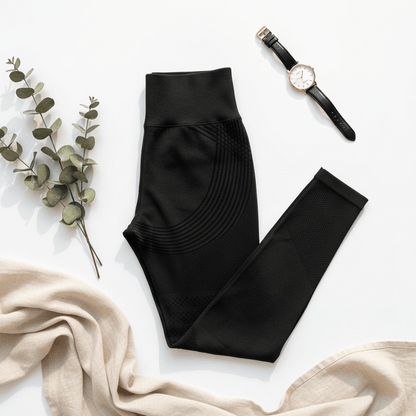Leggings noirs de sport pliés, motif de couture circulaire sur les hanches, posé à côté d’un bracelet montre et d’un drapé en rose clair avec des branches de eucalyptus. Pack Positiflow : Protocole Peau Lisse & Tonique.