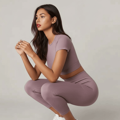 Femme en tenue de sport rose pâle, crop top court et leggings, accroupie et regardant loin, pose dynamique fitness.