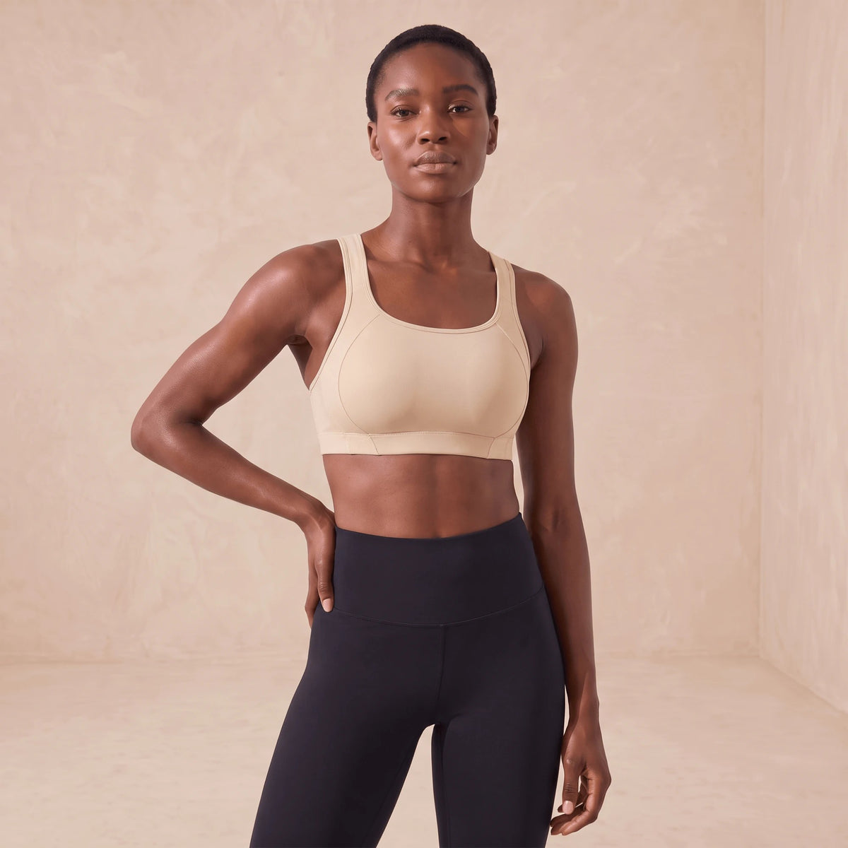Brassière de sport beige à maintien élevé, portée de face, mains sur les hanches, encolure ronde et bretelles larges