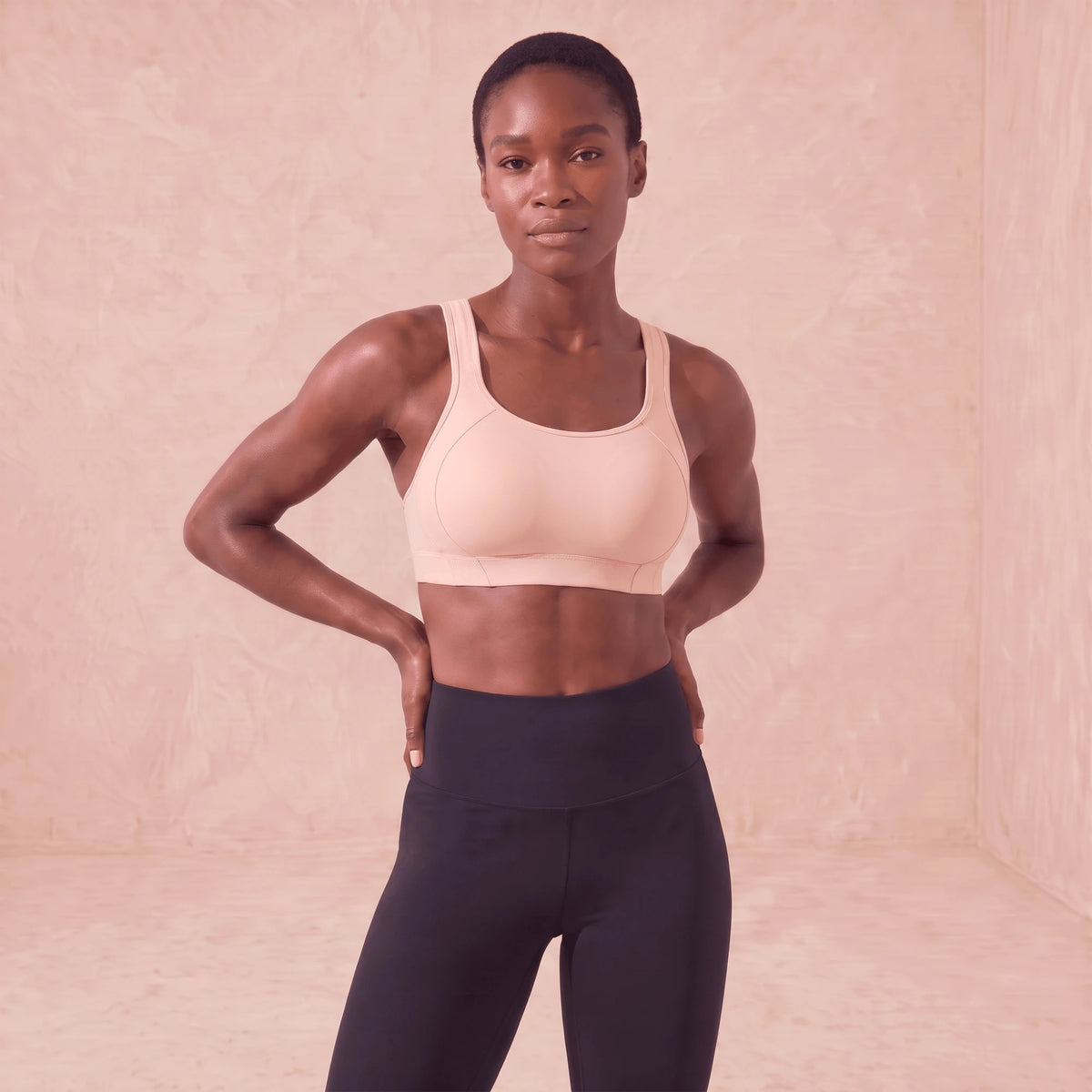 Brassière de sport rose clair à maintien élevé, portée de face, mains sur les hanches