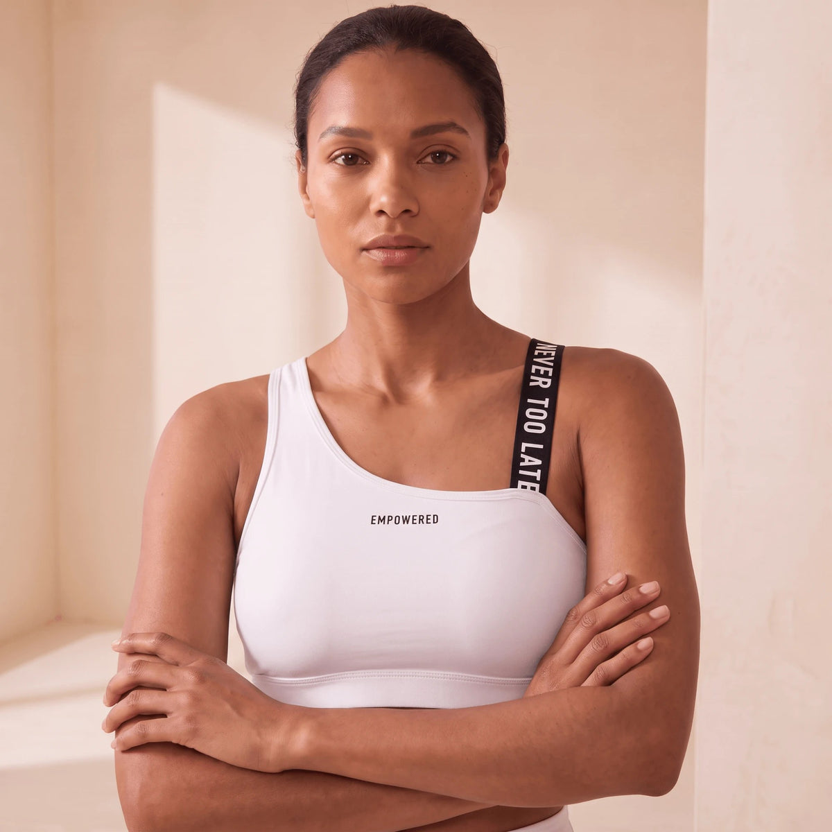 Brassière de sport blanche de face, bras croisés, bretelle à message noire et logo EMPOWERED discret