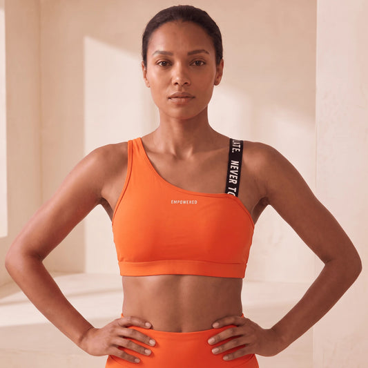 Brassière fitness orange portée de face, mains sur les hanches, bretelle à message noire et logo EMPOWERED sur la poitrine