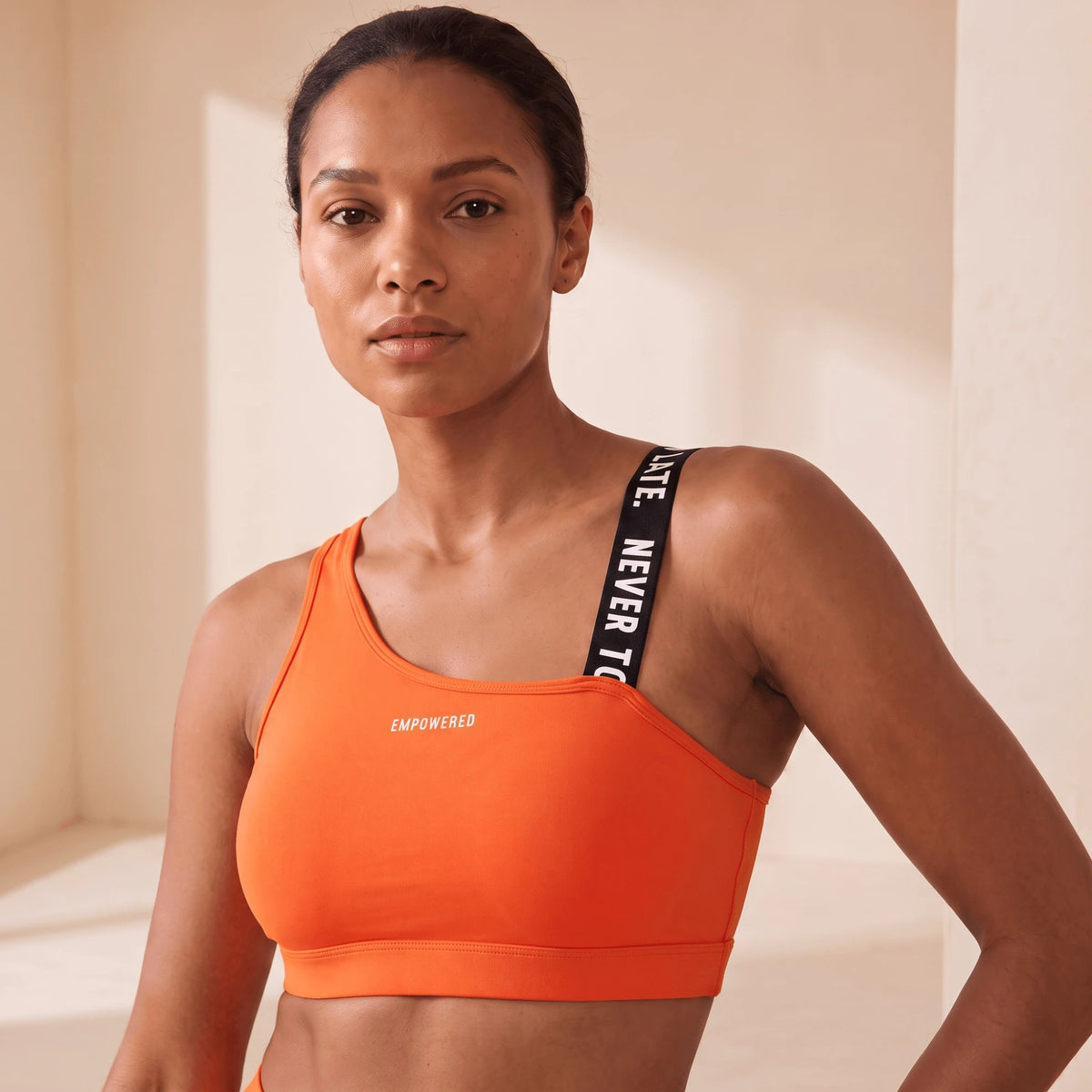 Brassière de sport orange vue de trois quarts, bretelle à message NEVER TOO LATE sur l'épaule
