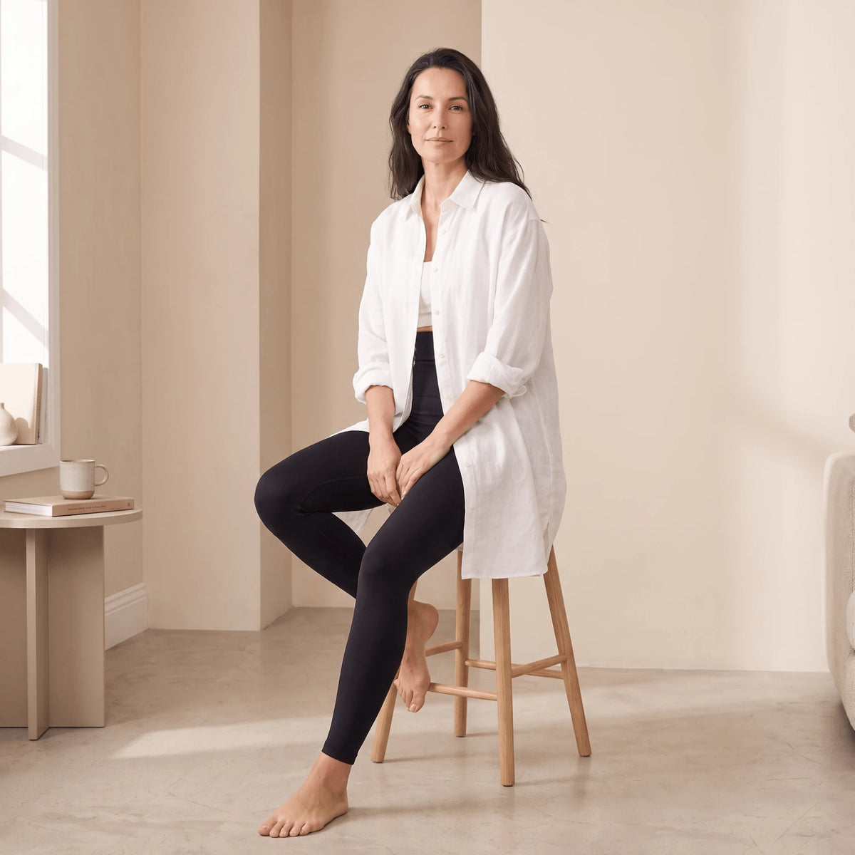 Collant gainant noir taille haute porté avec une chemise blanche longue, assise sur un tabouret en bois clair