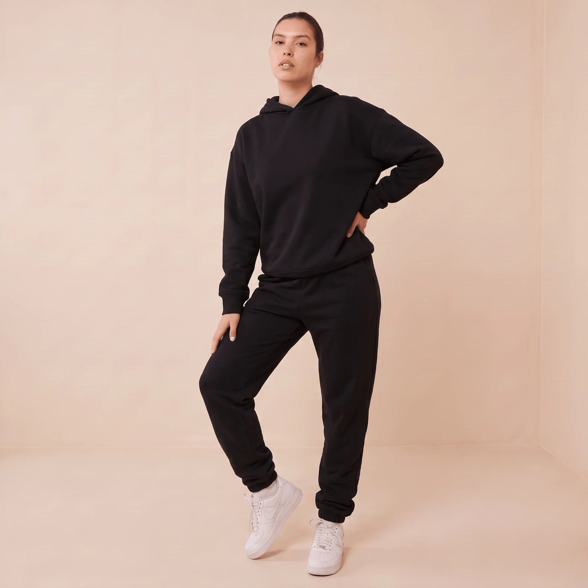 Ensemble de jogging femme noir : sweat à capuche et pantalon de jogging à bas élastiqué, portés avec des baskets blanches