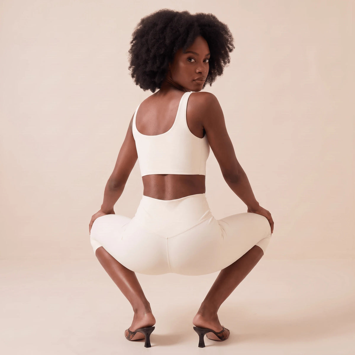 Legging blanc dos en V en squat de dos, regard par-dessus l'épaule : maille très élastique et effet pêche qui galbe visuellement