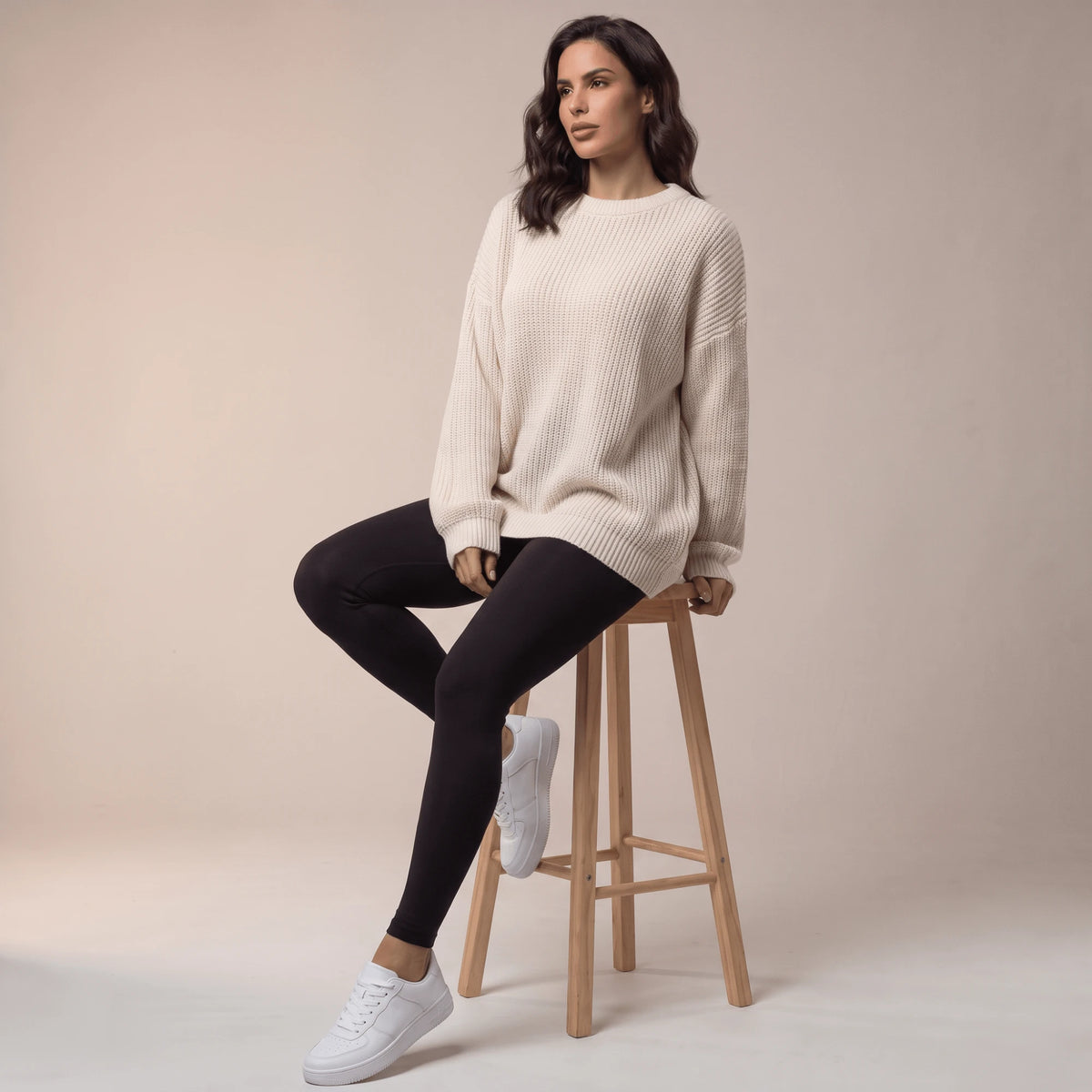 Legging de compression femme noir porté avec un grand pull crème, assise sur un tabouret en bois clair