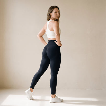Legging fitness femme noir taille haute vu de dos en trois quarts, brassière blanche, regard par-dessus l'épaule, studio crème