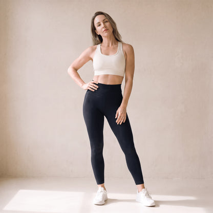 Legging fitness femme noir taille haute porté de face, une main sur la hanche, longueur cheville, studio crème