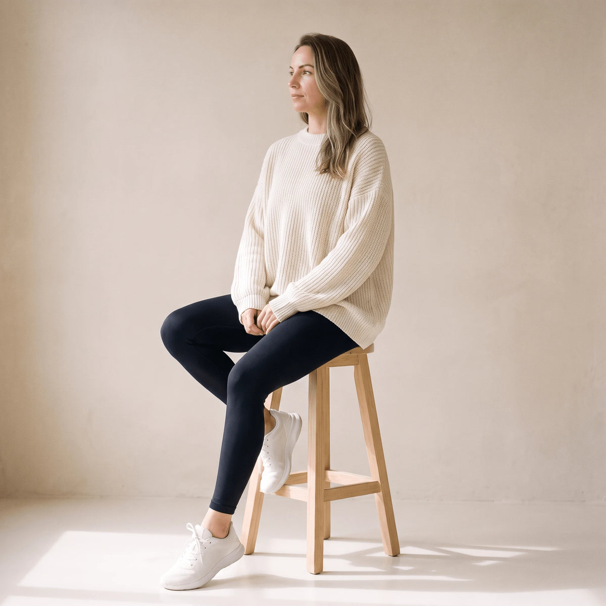 Legging fitness femme noir porté avec un grand pull crème, assise sur un tabouret en bois clair