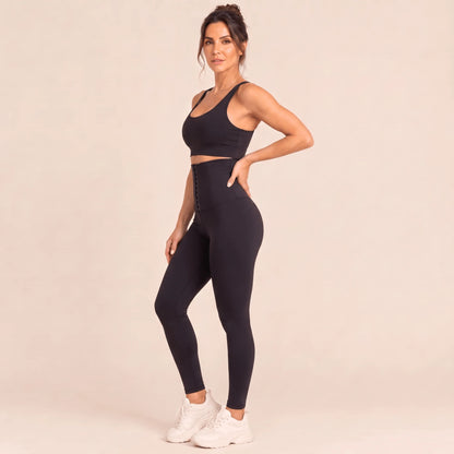 Legging sport femme gainant noir en trois-quarts, main dans le dos : ceinture haute à rangées de crochets et fessier galbé