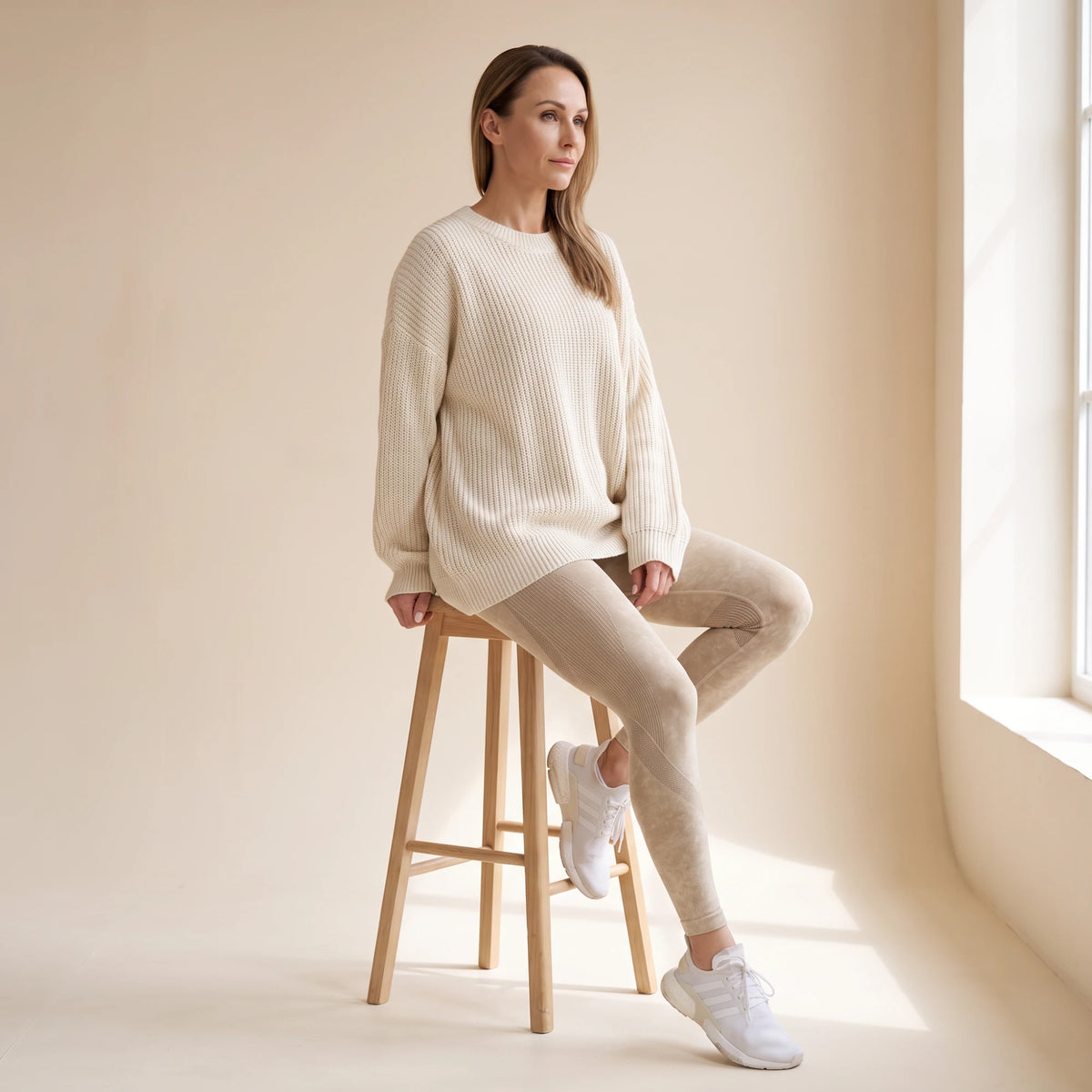 Legging galbant sans couture beige porté avec un grand pull crème, assise sur un tabouret en bois clair
