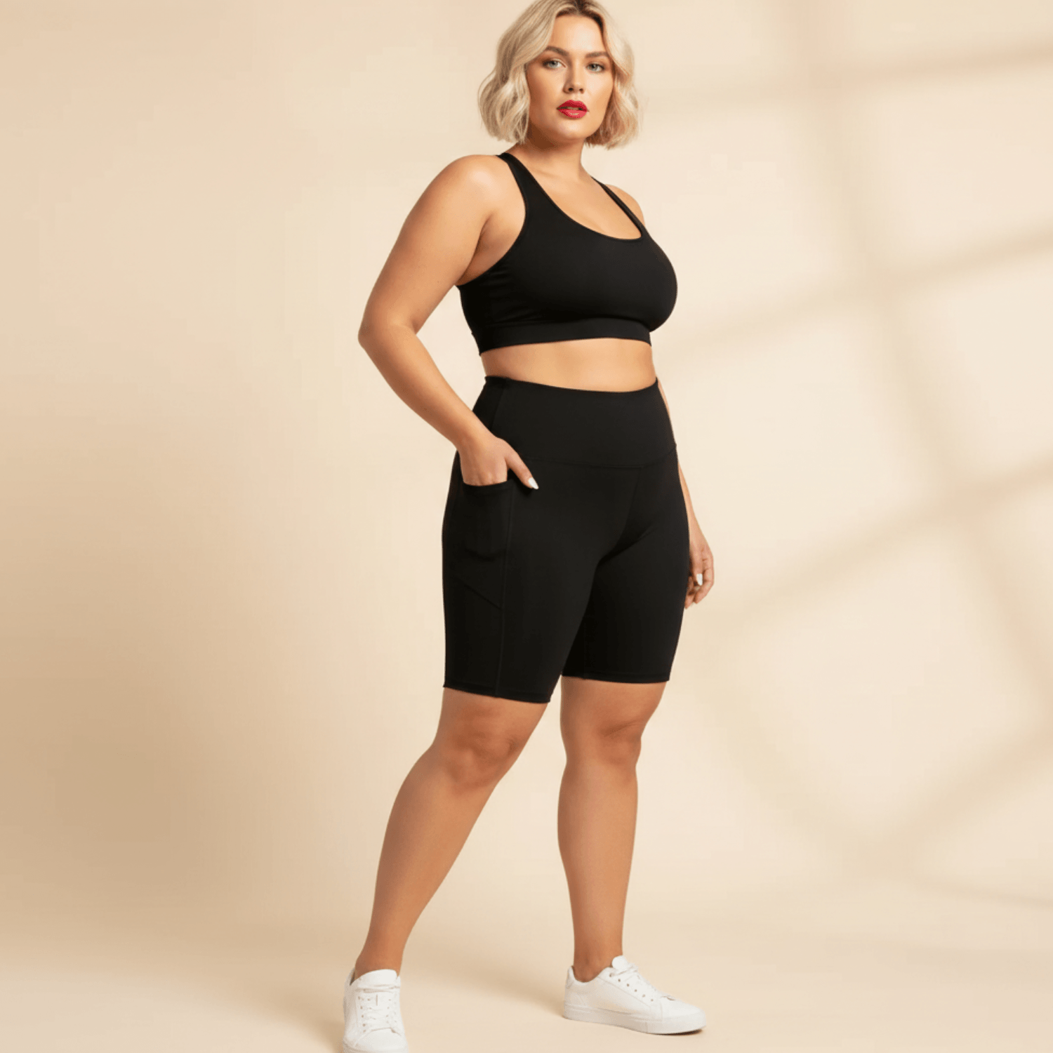 Short de sport femme grande taille noir cycliste taille haute avec poches latérales, vue de face trois-quarts, main dans la poche, fond studio crème