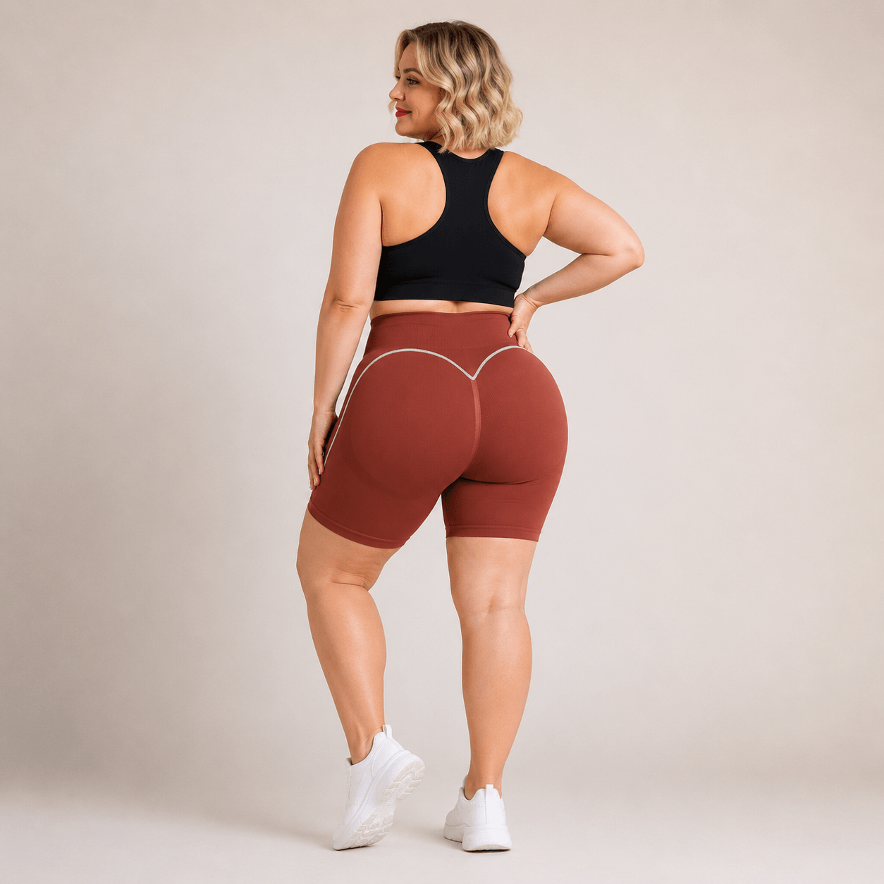 Short sport femme grande taille seamless terracotta, taille haute et liserés blancs en courbes, vue de dos avec effet galbant, visage tourné vers la caméra.