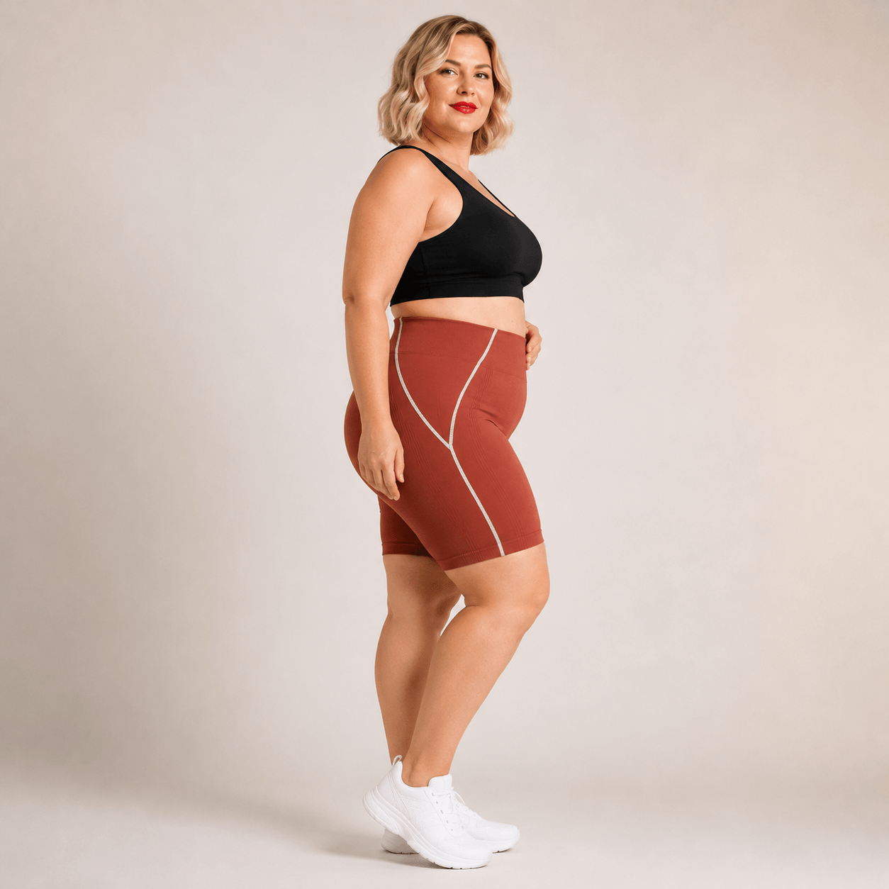 Short sport femme grande taille seamless terracotta, liserés blancs latéraux visibles, vue de profil visage vers la caméra, coupe gainante mi-cuisse.