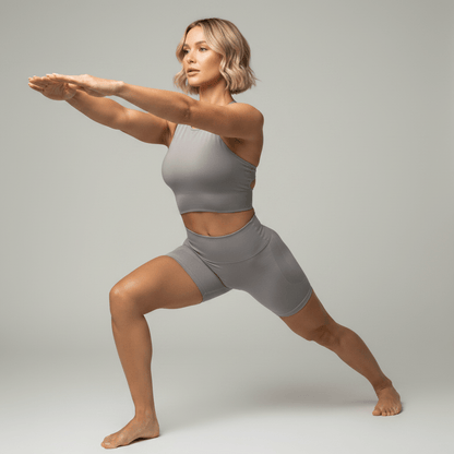 Brassière yoga hybride taupe, pose fente yoga dynamique bras étendus, portée avec short gris, studio minimaliste gris clair.