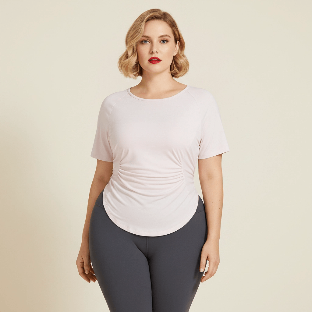 Top de yoga rose pâle pour femme grande taille, coupe cintrée avec ourlet arrondi et détails froncés, porté par une femme blonde avec un carré wavy dans un studio minimaliste.