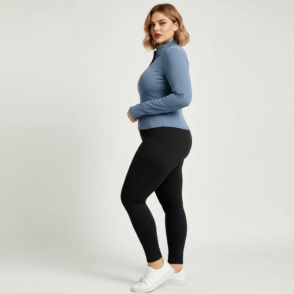 Veste de sport grande taille femme bleu-gris zippée, col montant, coupe ajustée cintrée, vue de profil avec mannequin visible, portée avec legging noir en studio fond crème.